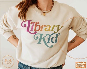 May include: A beige sweatshirt with the words "Library Kid" in colorful, retro-style lettering. The text is in shades of pink, orange, yellow, and blue. The sweatshirt has long sleeves and a crew neck.