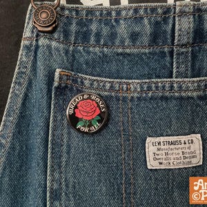 May include: Close-up of denim overalls with a "Bread & Roses For All" pin featuring a red rose. A Levi Strauss & Co. label is visible. The overalls have a metal clasp and are made of blue denim.