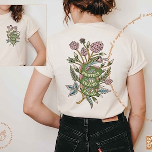 May include: White t-shirt with a green snake wrapped around pink flowers and leaves. The snake has the words "Protect Trans Folks" written on it. The shirt is a part of a collection where a portion of every sale is donated to causes we fight for.