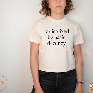 May include: Cream-colored t-shirt with the phrase "radicalized by basic decency" in black lettering. The shirt is paired with black jeans. The image also includes a logo and text in the lower right corner.