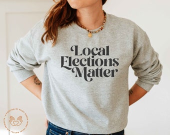 May include: A gray sweatshirt with the text "Local Elections Matter" printed in black on the front. The sweatshirt is being worn by a person with long brown hair.