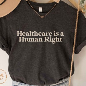 May include: A dark grey t-shirt with the text "Healthcare is a Human Right" printed in white. A portion of every sale is donated to causes we fight for.