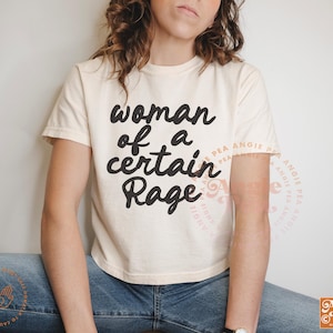 May include: Cream-coloured t-shirt featuring the phrase "woman of a certain Rage" in black script. The tee has short sleeves and a crew neck. The design is centered on the front of the shirt.