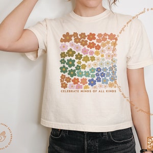 May include: A cream-colored cropped t-shirt with a graphic of colorful flowers arranged in a rainbow pattern. The text "Celebrate Minds of All Kinds" is printed below the flowers.