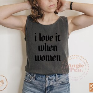 Op de afbeelding: Een donkergrijze tanktop met de tekst "i love it when women" in een gotische lettertype. De tanktop wordt gecombineerd met een blauwe spijkerbroek. De afbeelding bevat het Angie Pea-logo.