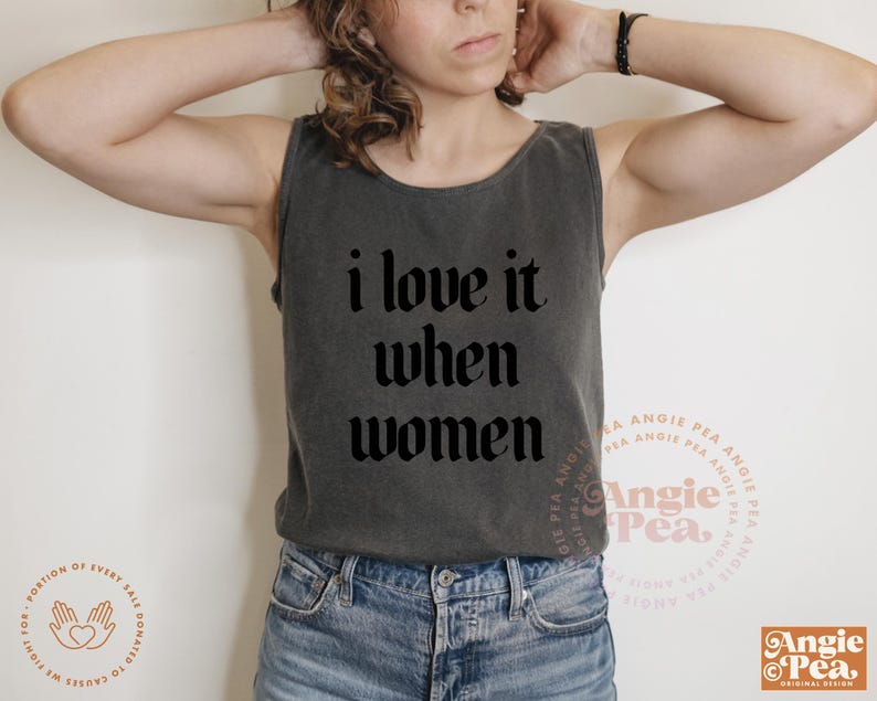 Op de afbeelding: Een donkergrijze tanktop met de tekst "i love it when women" in een gotische lettertype. De tanktop wordt gecombineerd met een blauwe spijkerbroek. De afbeelding bevat het Angie Pea-logo.