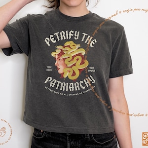 May include: A dark gray cropped t-shirt with a graphic of a woman's face with snakes for hair. The text "Petrify the Patriarchy" is printed on the shirt. The text "Take Back Our Power" is printed below the graphic. The text "Destruction to all systems of oppression" is printed below the text "Patriarchy".
