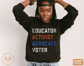May include: Black sweatshirt with the words "Educator, Activist, Advocate, Voter" in white, red, blue, and gray lettering. The sweatshirt is being worn by a person with dark skin and braids. The person is wearing a black baseball cap with a silver buckle.