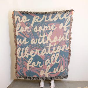 May include: A woven throw blanket with a pink and blue marbled pattern. The text "no pride for some of us without liberation for all of us" is printed in white on the blanket.