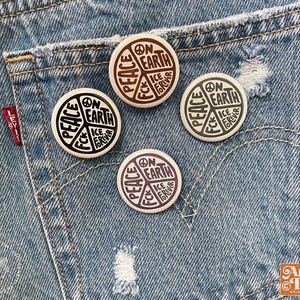 May include: Four round buttons with a peace symbol design, featuring the words "Peace," "On Earth," and "Ice Fck Forever." The buttons are in shades of black, brown, and purple, set against a denim background.
