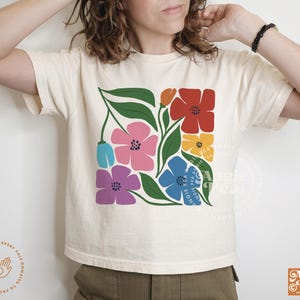 May include: Cream-colored t-shirt featuring a colorful floral design. The design includes flowers in red, pink, blue, yellow, and purple, with green leaves. The shirt has the text "Angie Pea" and a logo in the bottom right corner.
