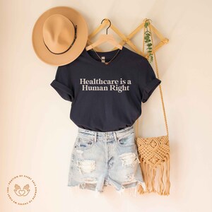 May include: A navy blue t-shirt with the text "Healthcare is a Human Right" in white lettering. The shirt is hanging on a wooden rack with a straw hat and a woven bag.