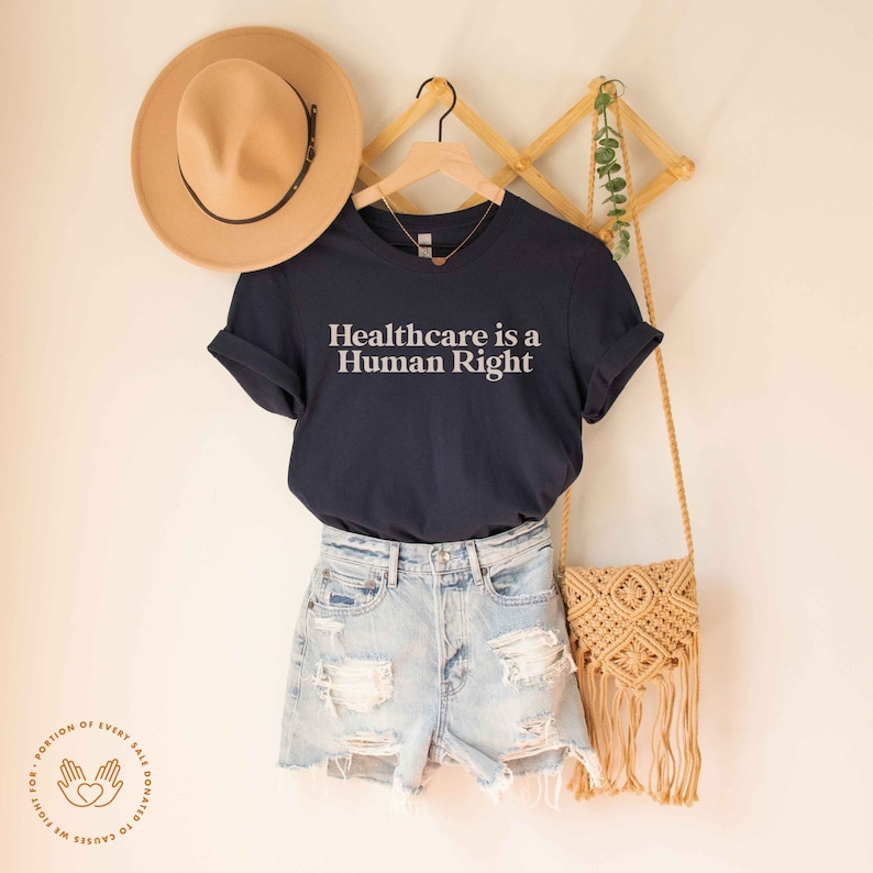 May include: A navy blue t-shirt with the text "Healthcare is a Human Right" in white lettering. The shirt is hanging on a wooden rack with a straw hat and a woven bag.