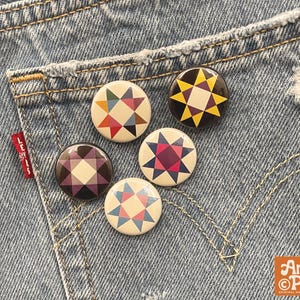 May include: Five circular pinback buttons featuring geometric star patterns in various color combinations including yellow, red, blue, purple, and cream. The buttons are arranged on a blue denim fabric surface with a red Levi's brand tag visible.