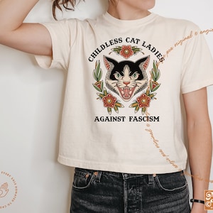 May include: A white t-shirt with a black and white cat illustration in a traditional tattoo style. The cat has a fierce expression and is surrounded by flowers. The text "Childless Cat Ladies Against Fascism" is printed on the shirt.