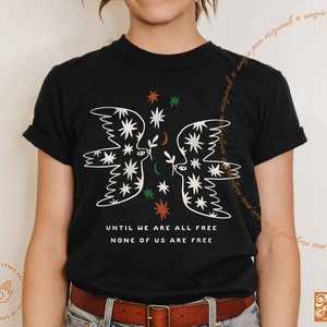 May include: Black t-shirt with a white graphic of two stylized birds facing each other. The graphic includes stars, moons, and leaves. The text "Until we are all free, none of us are free" is printed below the graphic.
