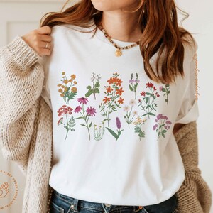 May include: White t-shirt with a floral print featuring a variety of flowers in shades of pink, red, orange, yellow, and purple.