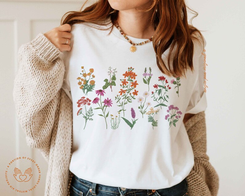 May include: White t-shirt with a floral print featuring a variety of flowers in shades of pink, red, orange, yellow, and purple.