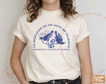 May include: A white t-shirt with a blue graphic of two birds on a branch. The graphic includes the text "La Migracion es un acto de Valentia" and "Migration is an act of courage".