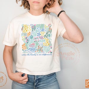 May include: Cream-colored t-shirt featuring a colorful floral design with the text "notice the beauty in our differences." The design includes various flowers in shades of yellow, blue, pink, and orange. The shirt has short sleeves and a crew neck.