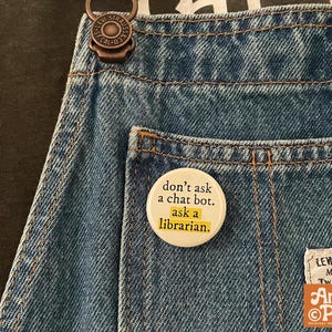 May include: Close-up of blue denim overalls with a round button pinned to the pocket. The button reads "don't ask a chat bot. ask a librarian." The overalls have a Levi Strauss tag.