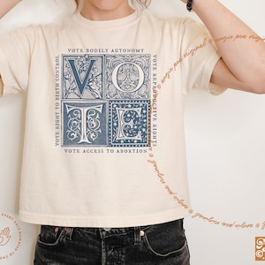 May include: A cream-colored t-shirt with a blue graphic of the word "VOTE" in a vintage style font. The graphic is made up of four squares, each with a different design. The text "VOTE RIGHT TO BIRTH CONTROL", "VOTE BODILY AUTONOMY", "VOTE REPRODUCTIVE RIGHTS", and "VOTE ACCESS TO ABORTION" surrounds the graphic.