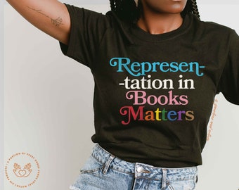 Könnte beinhalten: Ein schwarzes T-Shirt mit dem Text "Representation in Books Matters" in Regenbogenfarben. Der Text ist in einer stilisierten Schriftart.
