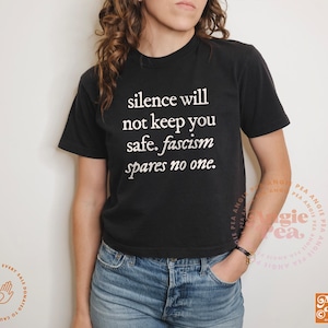 May include: Black t-shirt with the white text "silence will not keep you safe. fascism spares no one." The shirt is paired with blue denim jeans. The design is from Angie Pea Original Design.