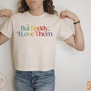 May include: A white cropped t-shirt with the text "But Daddy. I Love Them" printed in rainbow colors.