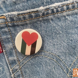 May include: A round button pin with a red heart and vertical stripes of green, brown, and black. The pin is set against a blue denim background. The pin is a design by Angie Pea.
