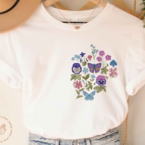 May include: White t-shirt with a floral design featuring purple, blue, and pink flowers and butterflies. The design is in the shape of a heart.