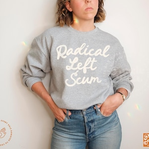 May include: A heather gray sweatshirt with the words "Radical Left Scum" in white script. The sweatshirt is worn with blue jeans. The image also includes the Angie Pea logo and a small graphic with text.