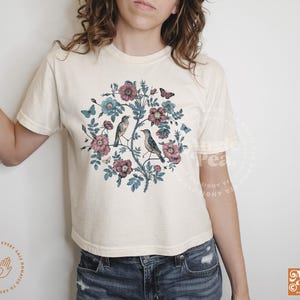 May include: A white t-shirt with a floral design featuring birds and butterflies. The design is in shades of blue, pink, and green.