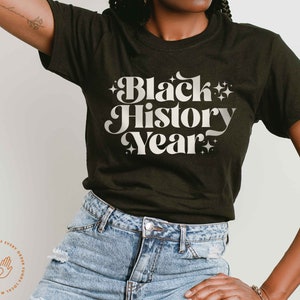 K&ouml;nnte beinhalten: Dunkelgr&uuml;nes T-Shirt mit dem Text "Black History Year" in wei&szlig;er Retro-Schrift. Der Text ist von Sternen umgeben. Das Model tr&auml;gt blaue Jeans.