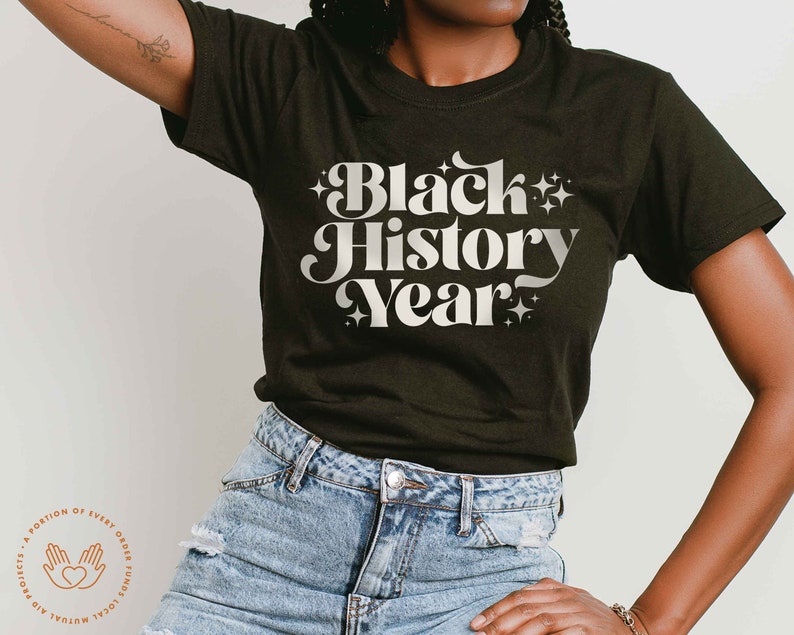 K&ouml;nnte beinhalten: Dunkelgr&uuml;nes T-Shirt mit dem Text "Black History Year" in wei&szlig;er Retro-Schrift. Der Text ist von Sternen umgeben. Das Model tr&auml;gt blaue Jeans.