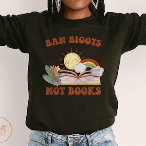 May include: Black sweatshirt with the text "Ban Bigots Not Books" in orange and white. The graphic features a book with a rainbow, clouds, and a sun with a smiling face.