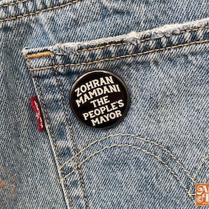 May include: A black button pinned to a blue denim pocket. The button features white text that reads "ZOHRAN MAMDANI THE PEOPLE'S MAYOR". The denim has a red Levi's tag.
