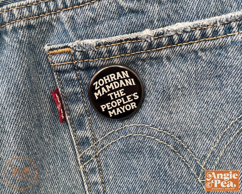 May include: A black button pinned to a blue denim pocket. The button features white text that reads "ZOHRAN MAMDANI THE PEOPLE'S MAYOR". The denim has a red Levi's tag.
