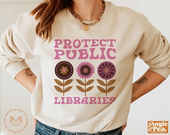 Op de afbeelding: Een beige sweatshirt met de tekst "Protect Public Libraries" in roze en bruine letters. De tekst wordt omringd door drie gestileerde roze en bruine bloemen met bruine stelen.