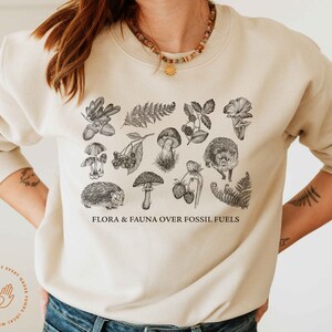 May include: A beige sweatshirt with a black and white illustration of flora and fauna. The text "FLORA & FAUNA OVER FOSSIL FUELS" is printed on the front of the sweatshirt.