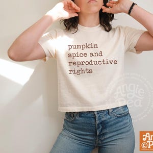 Puede incluir: Una camiseta de manga corta de color crema con el texto marrón "pumpkin spice and reproductive rights" impreso en la parte delantera. La camiseta tiene cuello redondo y dobladillo recto, combinada con pantalones vaqueros azules.