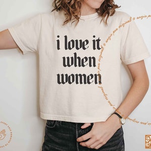 May include: Cream-colored t-shirt with the text "i love it when women" in a bold, black gothic font. The shirt features a cropped fit and short sleeves. The Angie Pea logo is visible in the bottom right corner.