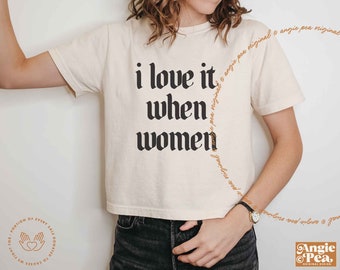 Op de afbeelding: Crèmekleurig T-shirt met de tekst "i love it when women" in een vet, zwart gotisch lettertype. Het shirt heeft een cropped pasvorm en korte mouwen. Het Angie Pea-logo is rechtsonder zichtbaar.