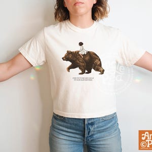 May include: Cream-colored t-shirt featuring a graphic of a bear carrying a person. The bear is brown, and the person is wearing white. The text "THE FUTURE BELONGS TO OUR DAUGHTERS" is below the graphic. The shirt has short sleeves.