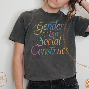 May include: A dark gray cropped t-shirt with the text "Gender is a Social Construct" in rainbow colors.