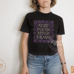 May include: Black t-shirt with the words "RESIST FASCISM REFUSE TYRANNY" in purple, surrounded by floral designs. The shirt is paired with blue jeans. The Angie Pea logo is visible on the lower right.