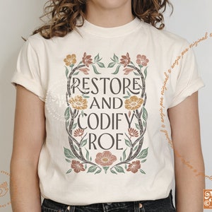 May include: A white t-shirt with a floral design and the text "Restore and Codify Roe" in brown and yellow lettering.