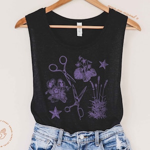 May include: Black tank top with a purple floral and scissor design. The design features a pair of scissors, flowers, and leaves.