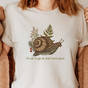 Op de afbeelding: Een wit T-shirt met een groene slakkenillustratie en de tekst "it's ok to go at your own pace".