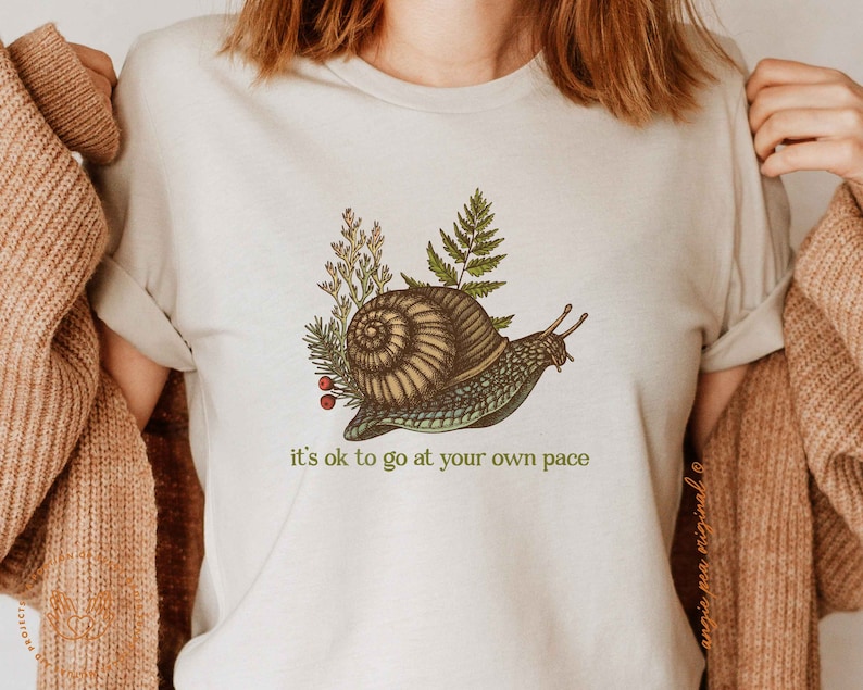 Op de afbeelding: Een wit T-shirt met een groene slakkenillustratie en de tekst "it's ok to go at your own pace".